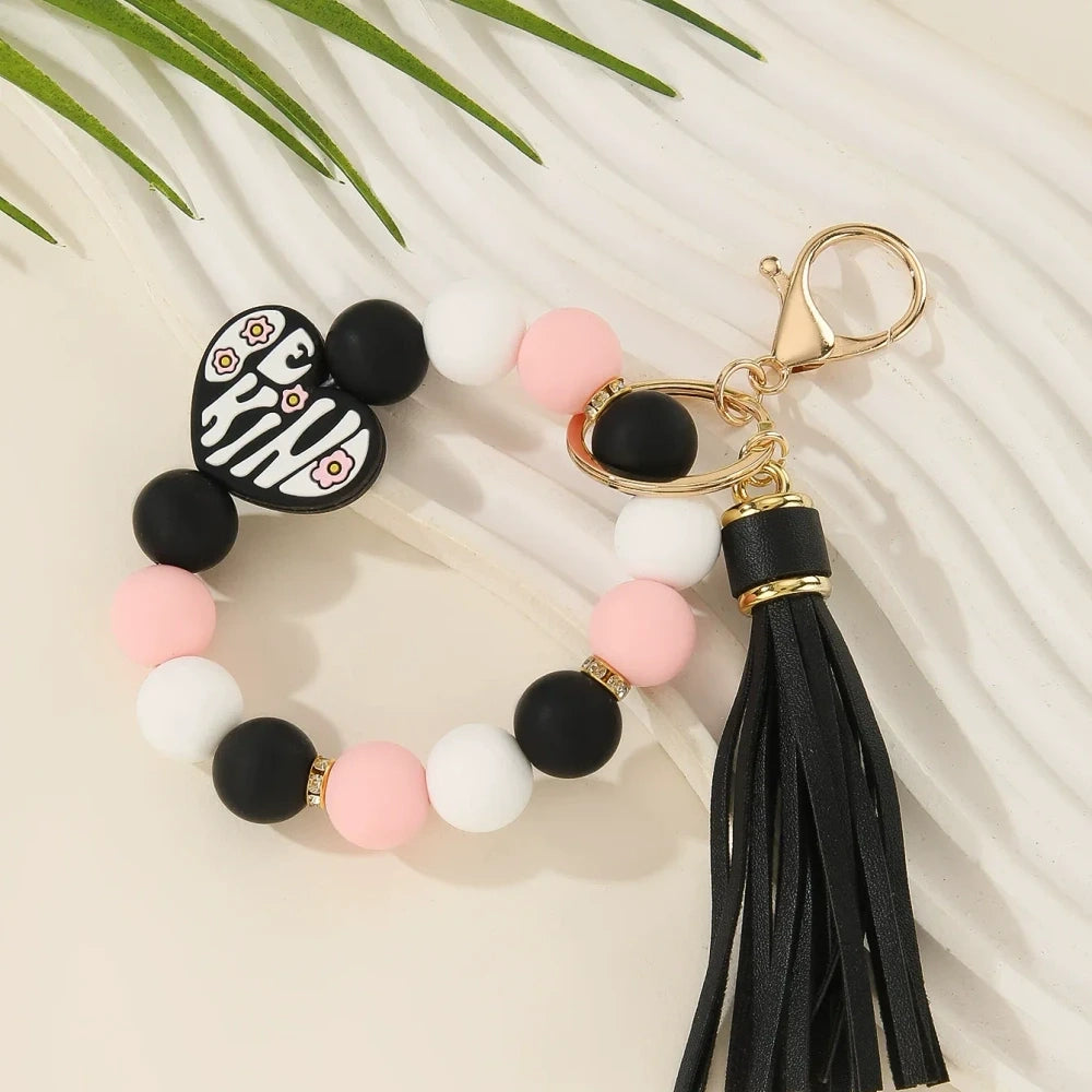 Be Kind heart printed silicone beads in pink, black, purple, and neutral tones shown on beaded wristlet keychains with tassels and gold hardware. - Black