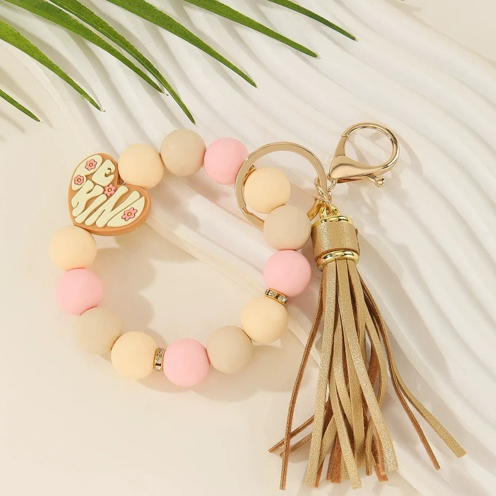 Be Kind heart printed silicone beads in pink, black, purple, and neutral tones shown on beaded wristlet keychains with tassels and gold hardware. - Brown