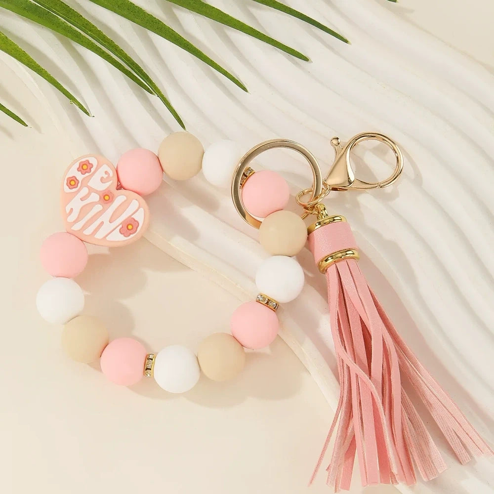Be Kind heart printed silicone beads in pink, black, purple, and neutral tones shown on beaded wristlet keychains with tassels and gold hardware. - Pink
