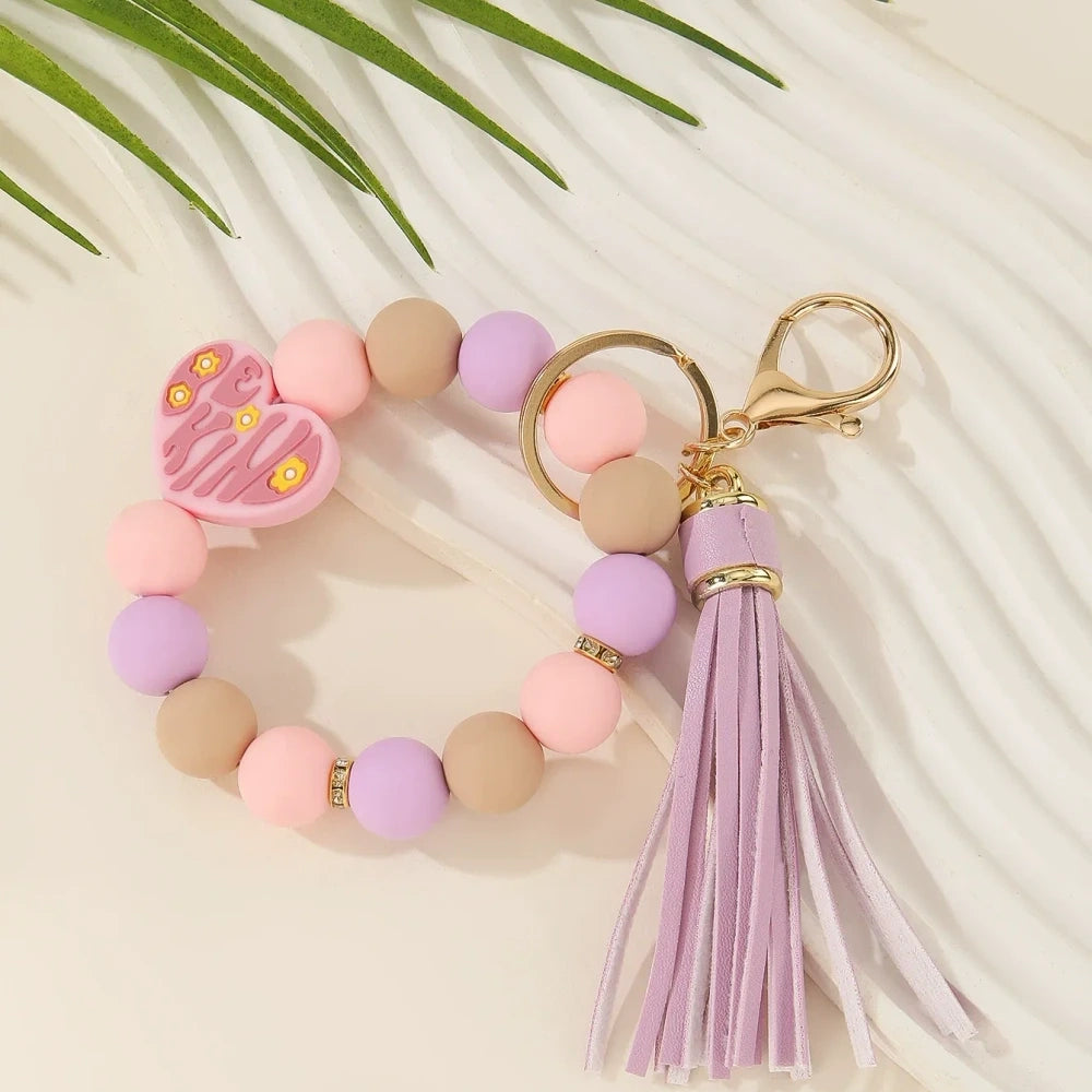 Be Kind heart printed silicone beads in pink, black, purple, and neutral tones shown on beaded wristlet keychains with tassels and gold hardware. - Purple