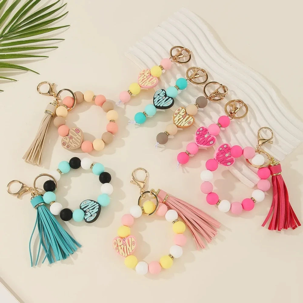 Colorful Be Kind heart printed silicone beads displayed on beaded bracelet keychains with tassels in pink, teal, yellow, and neutral tones on a cream background.