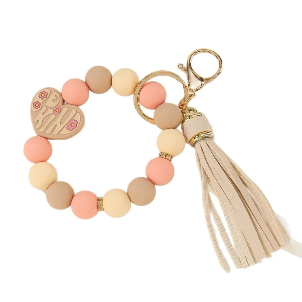 Colorful Be Kind heart printed silicone beads displayed on beaded bracelet keychains with tassels in pink, teal, yellow, and neutral tones on a cream background. - 1