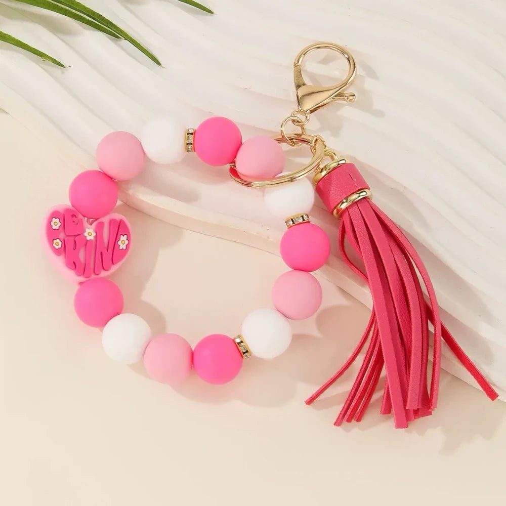 Colorful Be Kind heart printed silicone beads displayed on beaded bracelet keychains with tassels in pink, teal, yellow, and neutral tones on a cream background. - 2