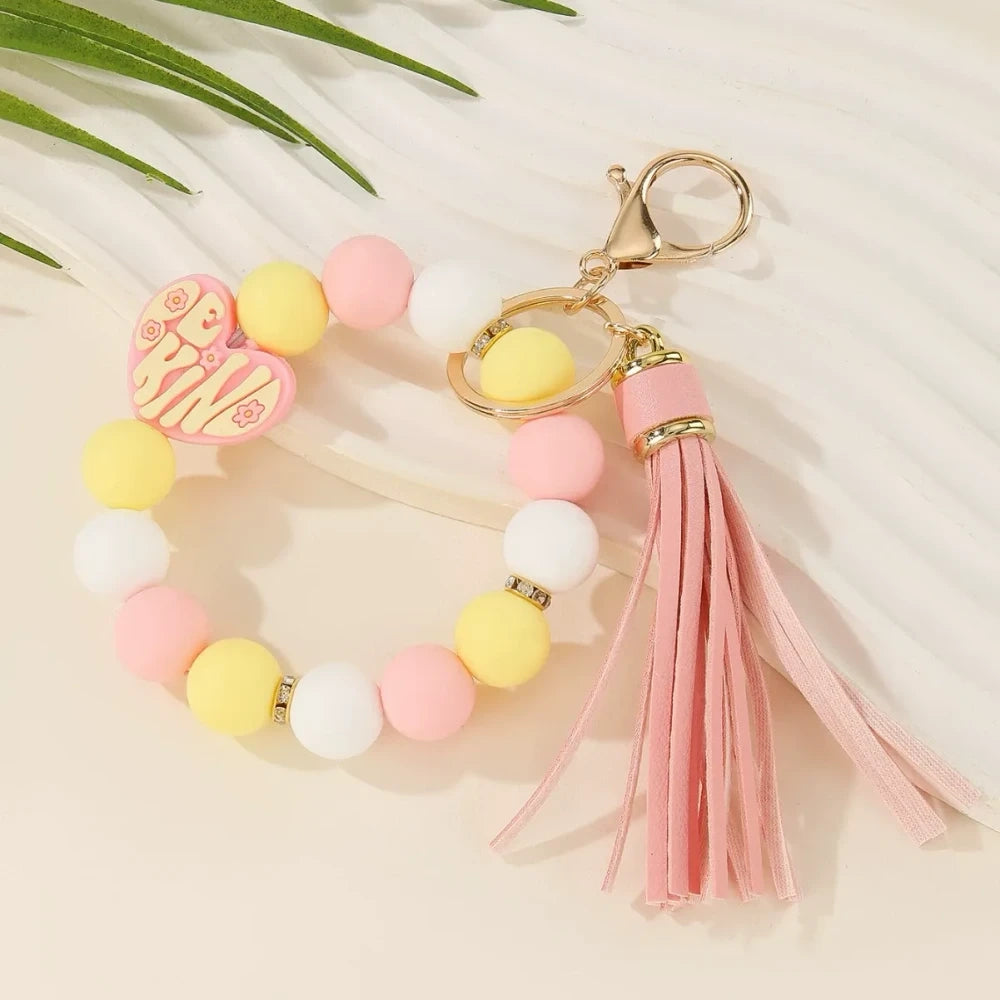 Colorful Be Kind heart printed silicone beads displayed on beaded bracelet keychains with tassels in pink, teal, yellow, and neutral tones on a cream background. - 3