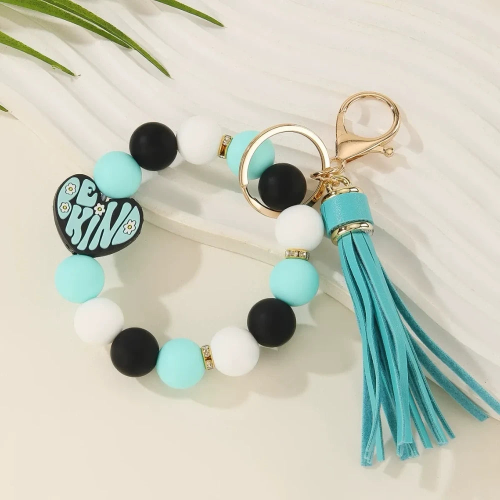 Colorful Be Kind heart printed silicone beads displayed on beaded bracelet keychains with tassels in pink, teal, yellow, and neutral tones on a cream background. - 4