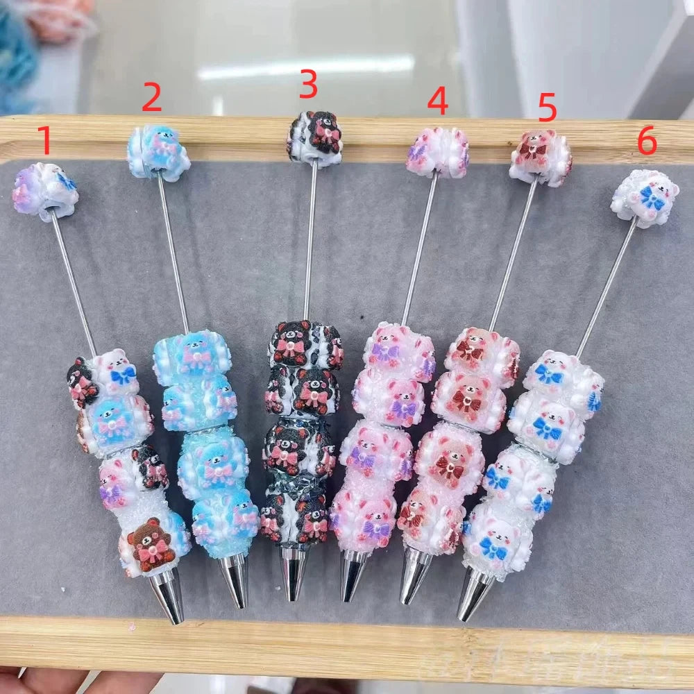 Six colorful clay beaded ballpoint pens featuring cute bear and bow resin beads in blue, black, pink, mauve, and white glitter styles, numbered 1 through 6, displayed on a wooden tray.