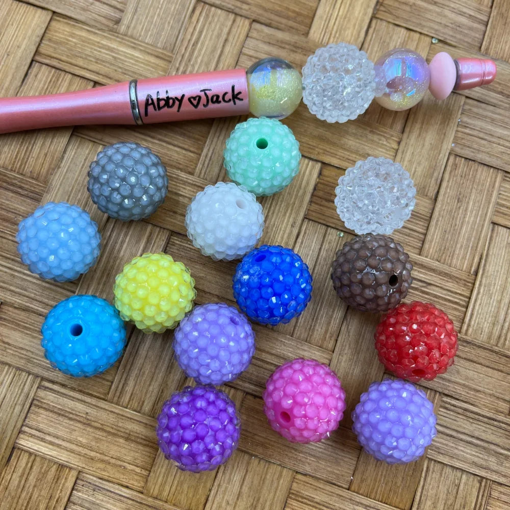 Assorted color berry bumpy rhinestone beads in gray, blue, teal, white, yellow, purple, pink, red, brown, and clear iridescent finishes displayed on wicker surface next to a beadable pen labeled Abby Jack.