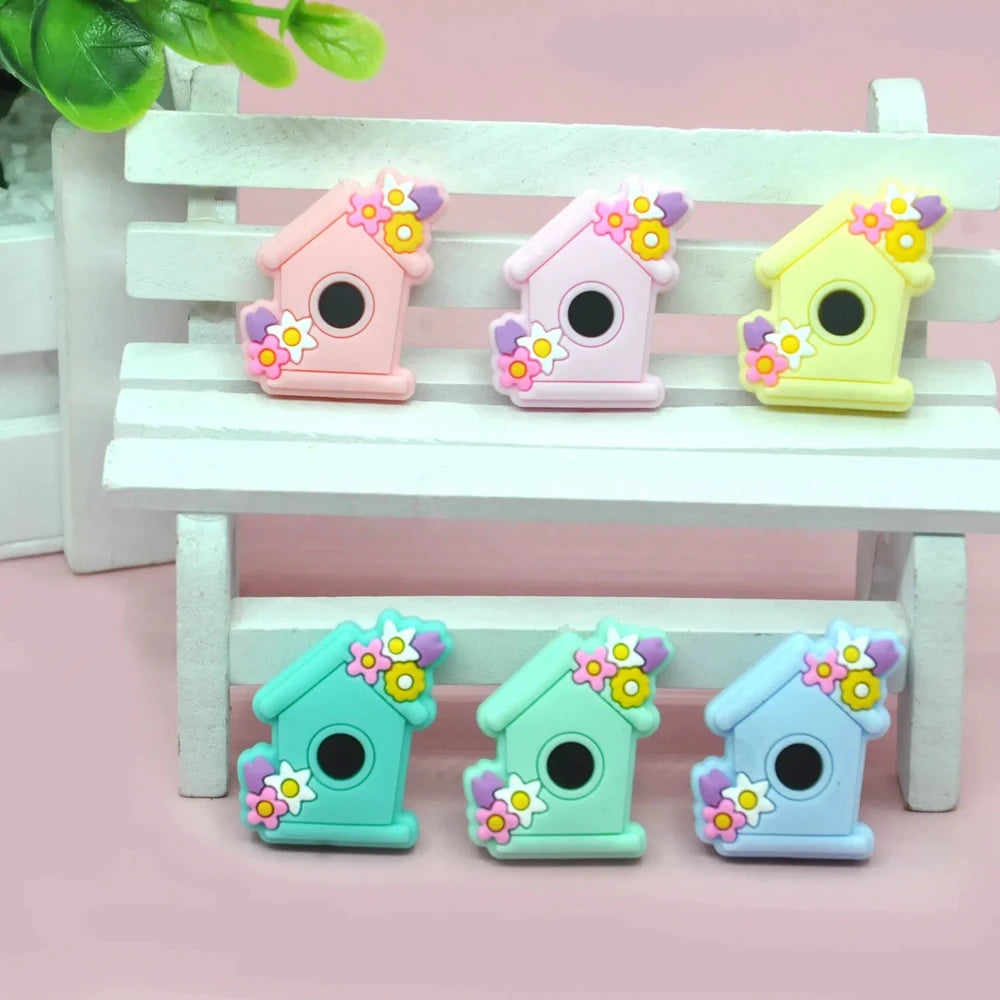 Six pastel silicone focal beads shaped like birdhouses decorated with flowers in pink, lavender, yellow, teal, mint, and blue colors displayed on a miniature white bench.