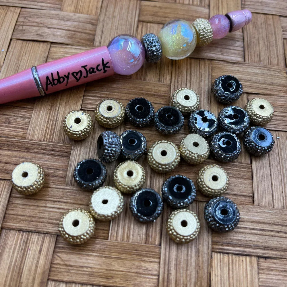 Black and gold textured acrylic spacer beads with dotted rim edges and center hole, shown next to a decorated beadable pen on a wicker surface.