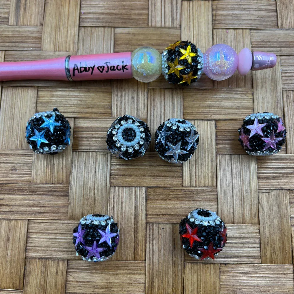 Black rhinestone chunky clay beads with colorful star embellishments in blue, yellow, pink, purple, red, and clear, displayed on a wicker surface with a decorated beadable pen.