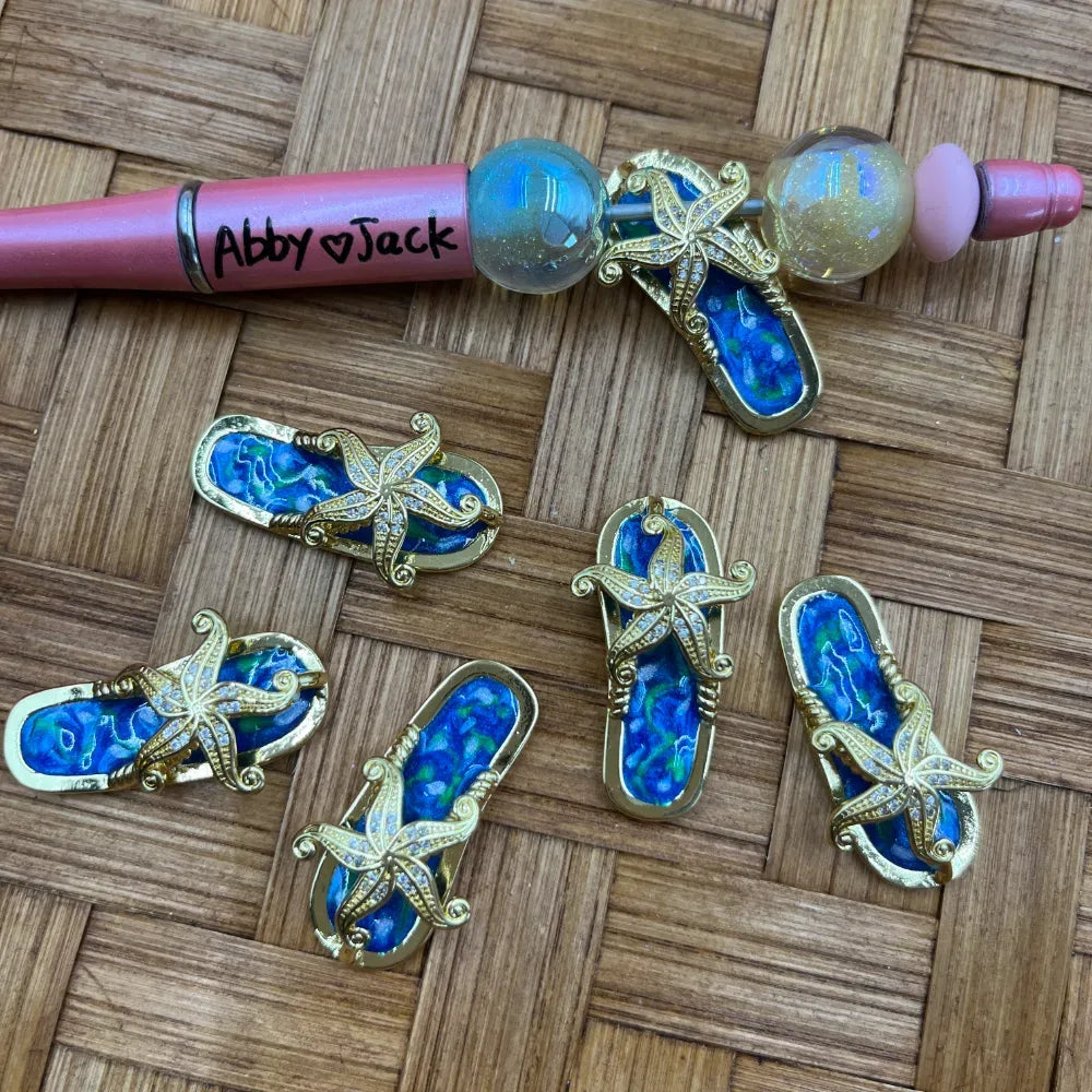 Blue and gold enamel starfish flip flop bail beads displayed next to a beadable pen on a wicker background, perfect for DIY jewelry making and beadable pens.