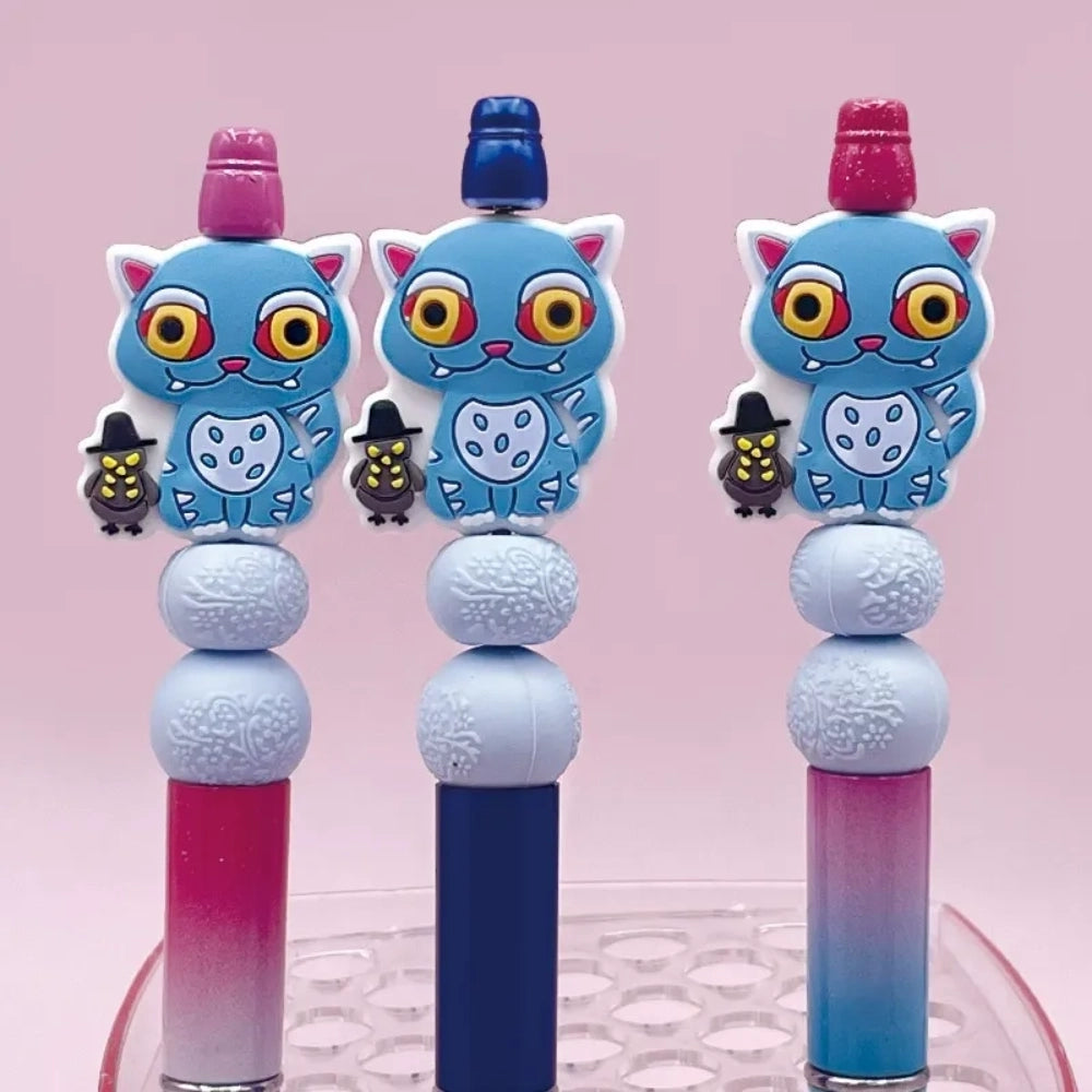 Blue monster cat silicone focal bead with large yellow eyes, fangs, and tiny witch owl charm, shown on beadable pens in pink, blue, and purple colorways on a pink background.