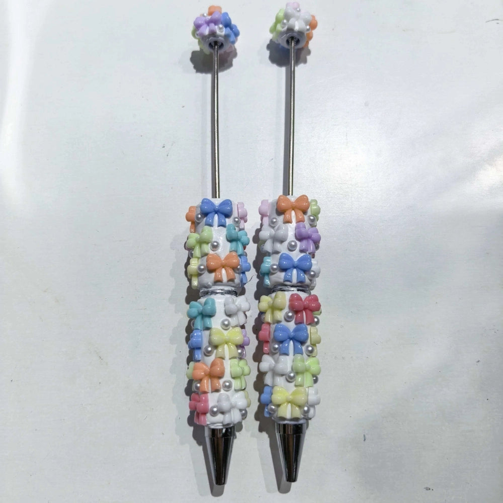 Two white clay beadable pens decorated with pastel multicolor bow and pearl embellishments, silver tip and topper, on white background.