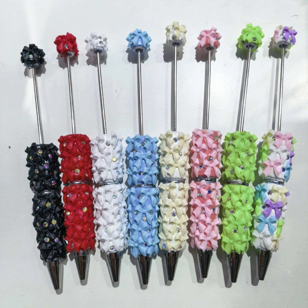 Eight decorative clay beadable pens covered in mini ribbon bows and rhinestone gems in colors including black, red, white, blue, cream, pink, green, and purple with silver metal tips and topper beads.