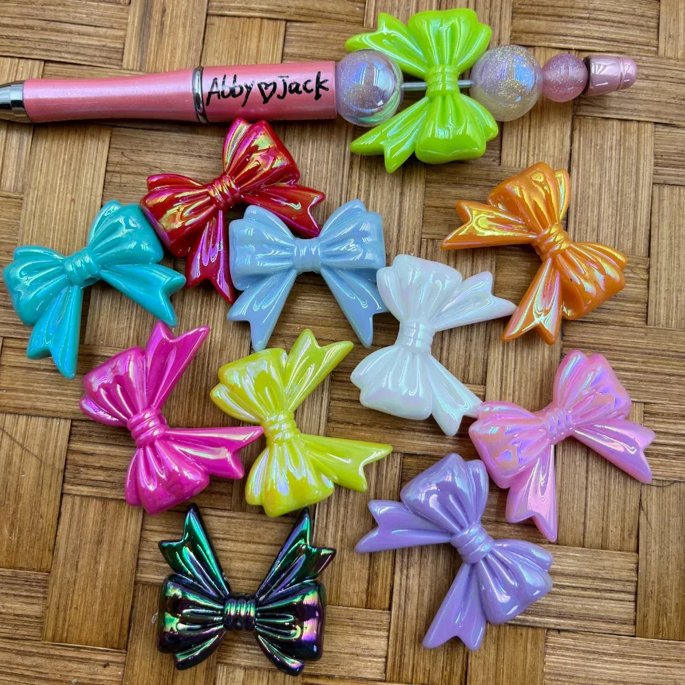 Assorted iridescent pearlescent bow-shaped acrylic beads in multiple colors including pink, teal, red, yellow, purple, orange, white, and holographic black, displayed on a woven surface with a beadable pen.
