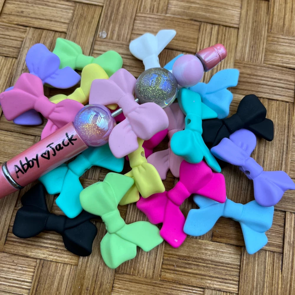 Colorful matte bow tie acrylic beads in assorted pastel and neon colors including pink, teal, purple, black, yellow, and white, displayed with beadable pens on a wicker surface.
