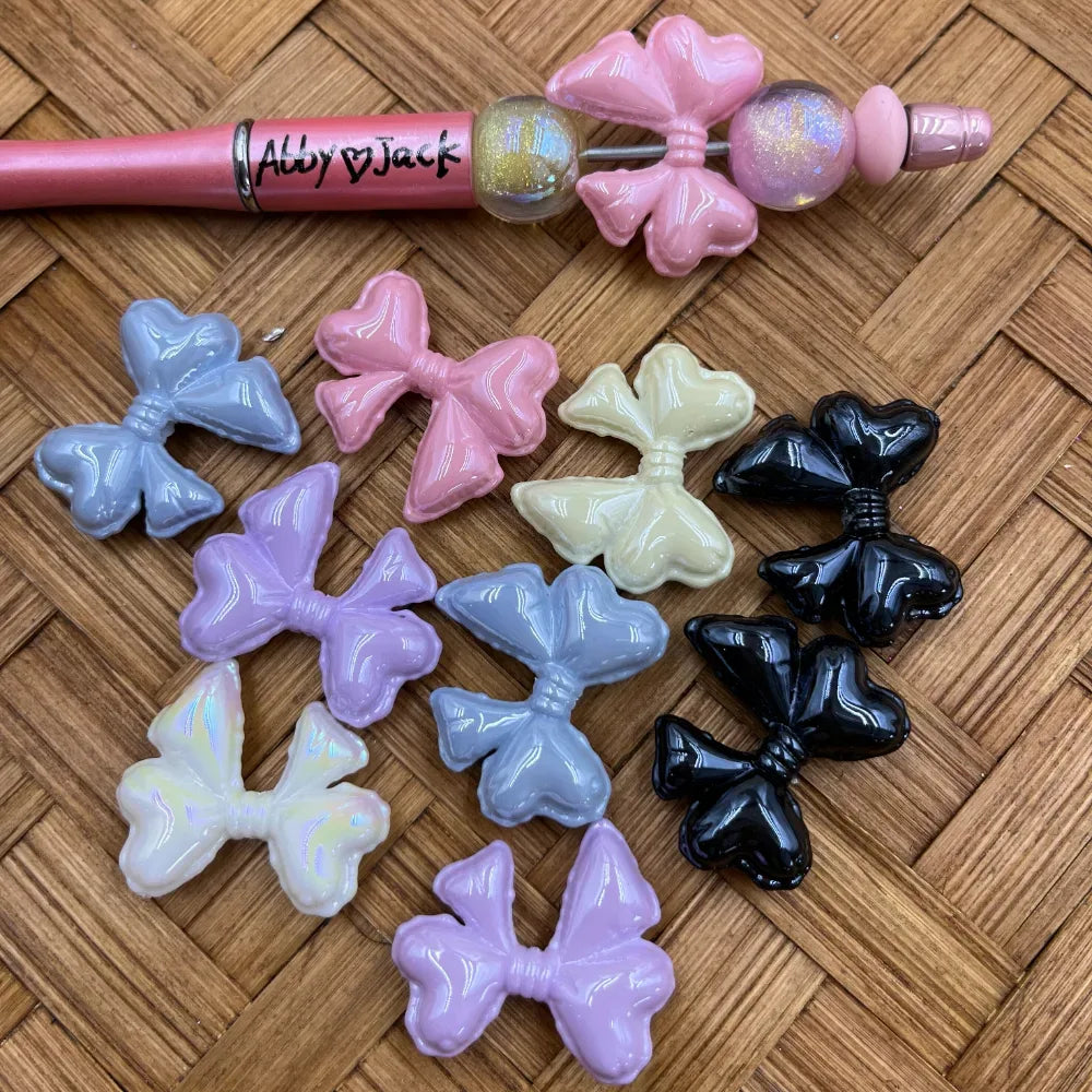 Assorted bow tie heart-shaped acrylic beads in pastel purple, pink, gray, cream, iridescent white, and black finishes displayed on a woven surface next to a pink beadable pen with bow bead accent.
