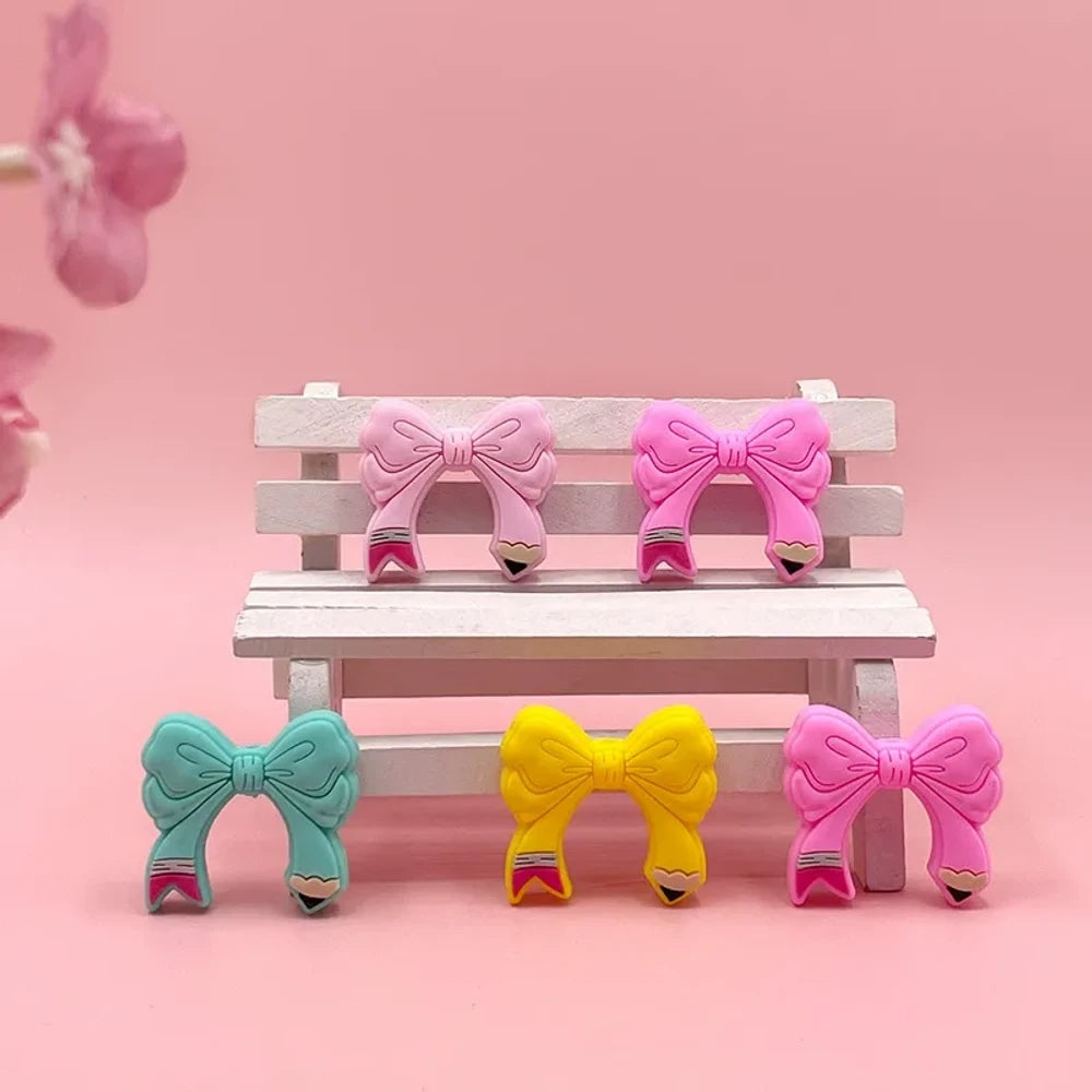 Assorted colorful bow tie silicone focal beads in light pink, hot pink, teal, yellow, and magenta displayed on a miniature white bench with pink background