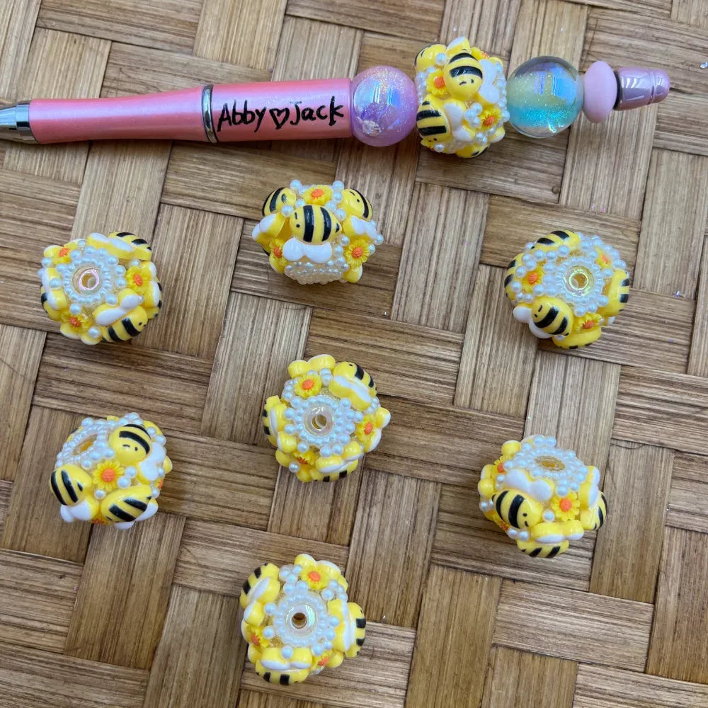 Bumble bee and daisy flower resin fancy beads with white pearl accents displayed on a woven surface next to a pink beadable pen decorated with matching bee beads.
