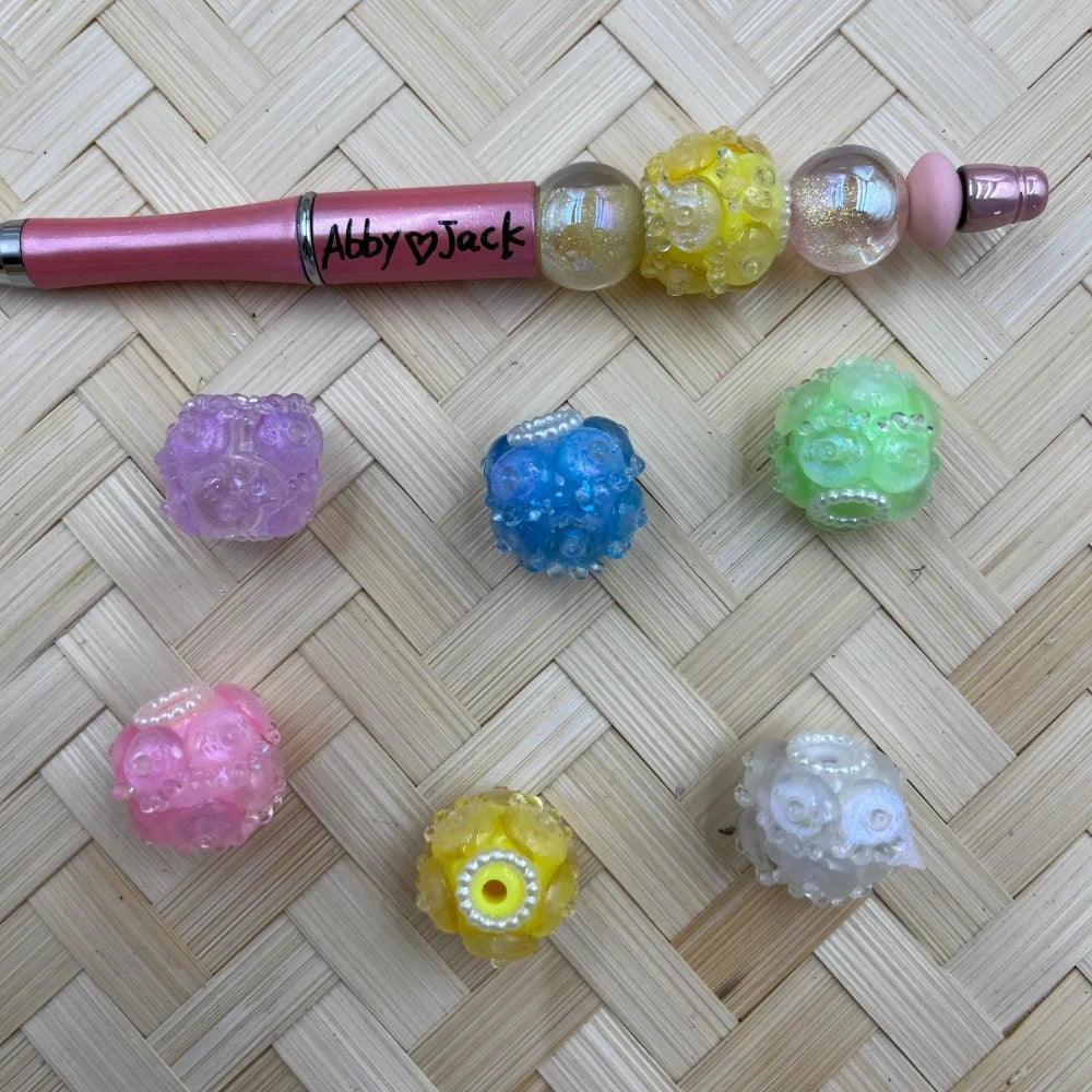 Assorted pastel bumpy bubble cluster resin fancy beads in purple, blue, green, pink, yellow, and white, displayed on a wicker surface alongside a pink beadable pen decorated with the same style beads.