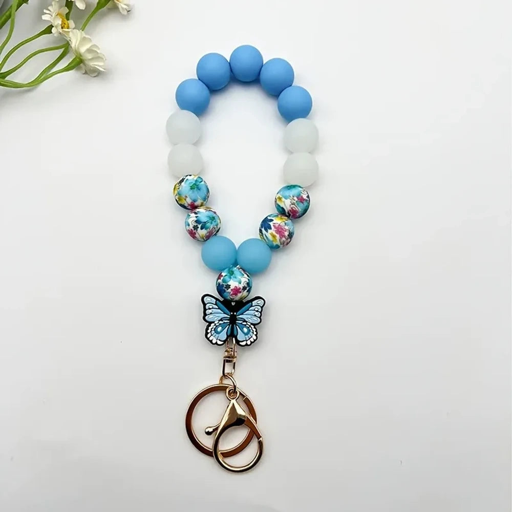 Assorted butterfly charm silicone bead bracelets arranged in a fan display featuring floral, leopard, rainbow, and solid color printed silicone beads with decorative butterfly pendants and daisy flowers at center. - Blue