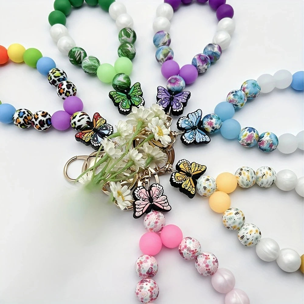 Assorted butterfly charm silicone bead bracelets arranged in a fan display featuring floral, leopard, rainbow, and solid color printed silicone beads with decorative butterfly pendants and daisy flowers at center.