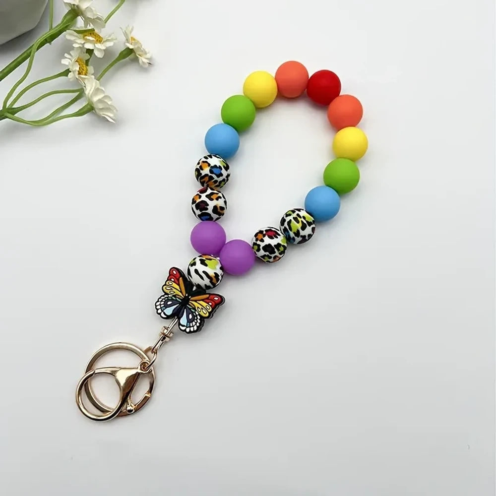 Assorted butterfly charm silicone bead bracelets arranged in a fan display featuring floral, leopard, rainbow, and solid color printed silicone beads with decorative butterfly pendants and daisy flowers at center. - Colorful