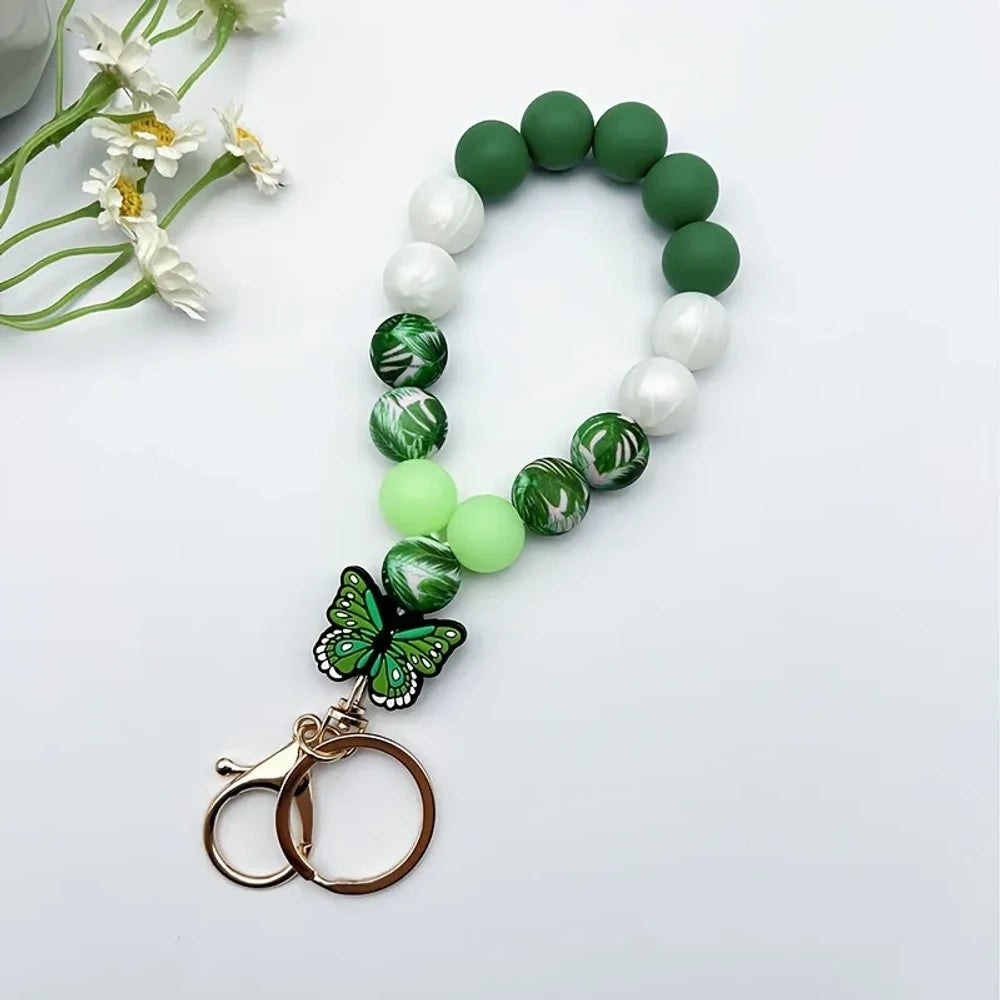 Assorted butterfly charm silicone bead bracelets arranged in a fan display featuring floral, leopard, rainbow, and solid color printed silicone beads with decorative butterfly pendants and daisy flowers at center. - Green