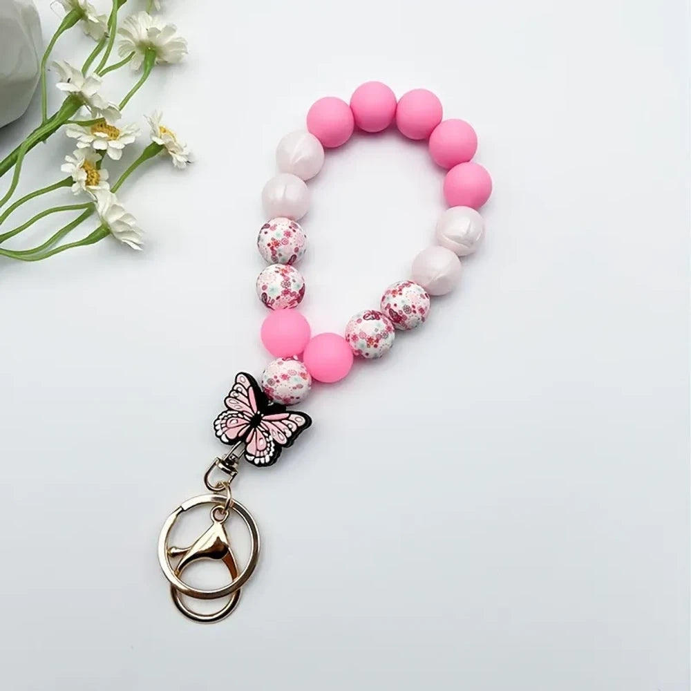 Assorted butterfly charm silicone bead bracelets arranged in a fan display featuring floral, leopard, rainbow, and solid color printed silicone beads with decorative butterfly pendants and daisy flowers at center. - Pink