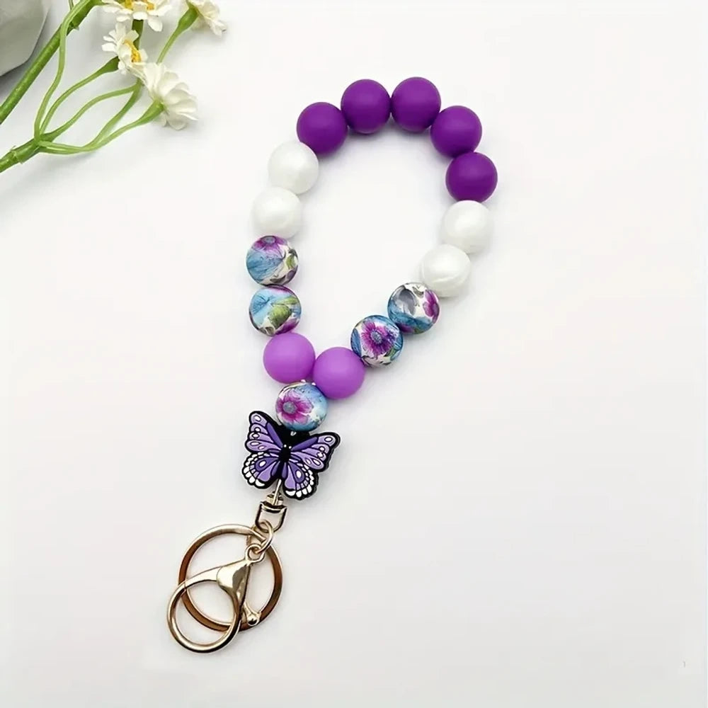 Assorted butterfly charm silicone bead bracelets arranged in a fan display featuring floral, leopard, rainbow, and solid color printed silicone beads with decorative butterfly pendants and daisy flowers at center. - Purple