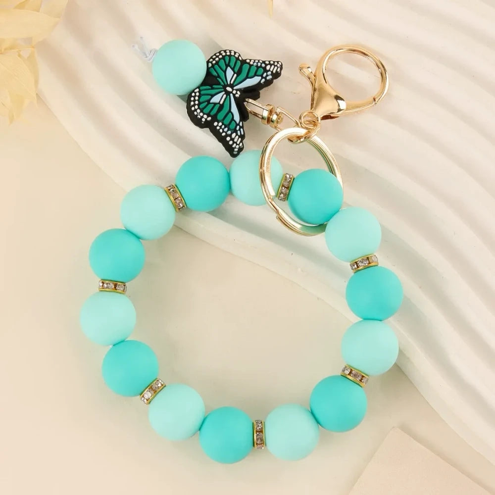Four silicone bead bracelet keychains with butterfly charms in pink, teal, red-yellow, and purple colorways with gold lobster clasp hardware on a cream background. - Blue