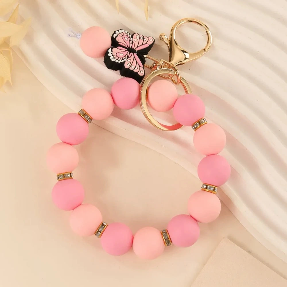 Four silicone bead bracelet keychains with butterfly charms in pink, teal, red-yellow, and purple colorways with gold lobster clasp hardware on a cream background. - Pink