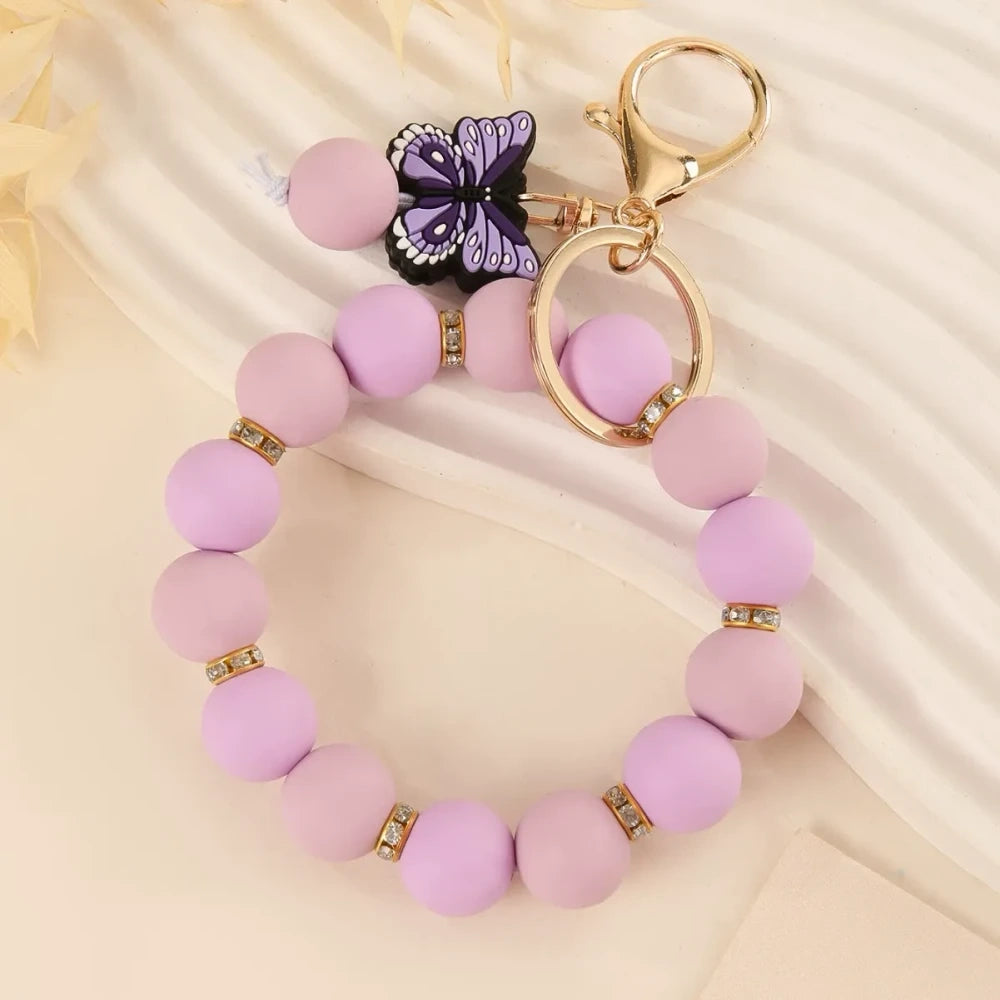 Four silicone bead bracelet keychains with butterfly charms in pink, teal, red-yellow, and purple colorways with gold lobster clasp hardware on a cream background. - Purple