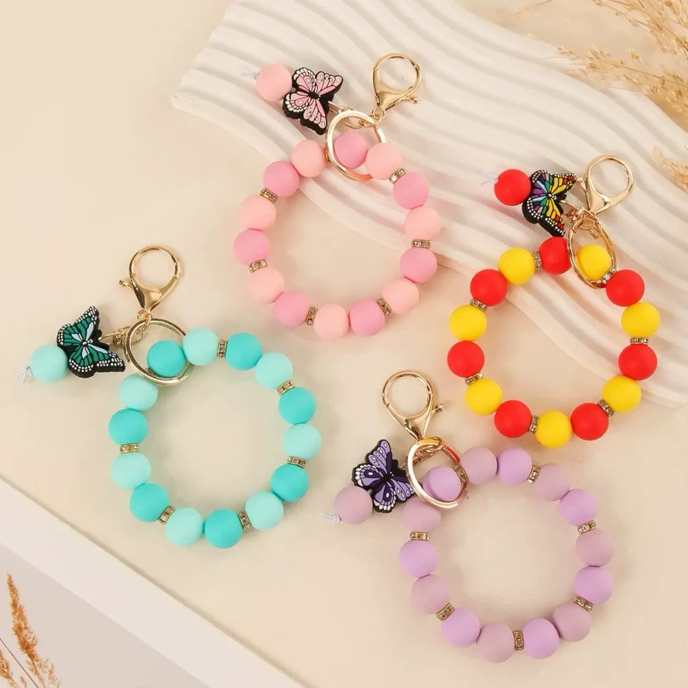 Four silicone bead bracelet keychains with butterfly charms in pink, teal, red-yellow, and purple colorways with gold lobster clasp hardware on a cream background. - Random Mixed