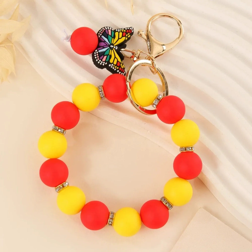 Four silicone bead bracelet keychains with butterfly charms in pink, teal, red-yellow, and purple colorways with gold lobster clasp hardware on a cream background. - Red