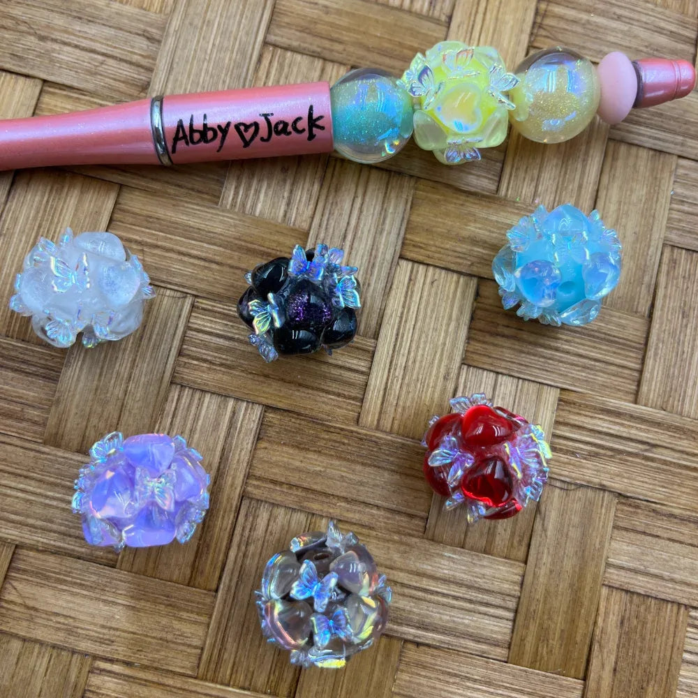Assorted iridescent butterfly cluster resin fancy beads in white, black, blue, yellow, purple, red, and clear colors displayed on a woven surface next to a pink beadable pen labeled Abby Jack.