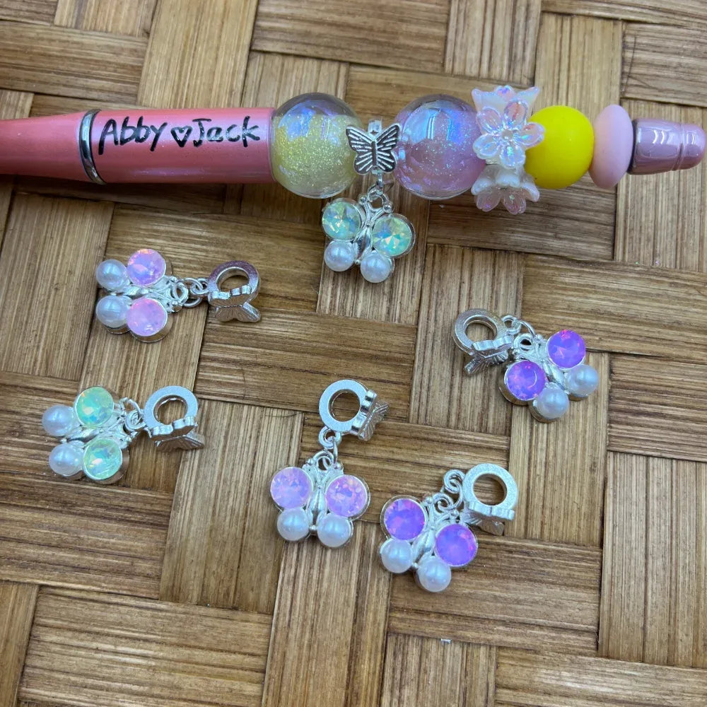 Silver butterfly charm bail beads with iridescent crystal and pearl accents in pink, purple, and white opal colors, displayed next to a decorated beadable pen on a woven surface.