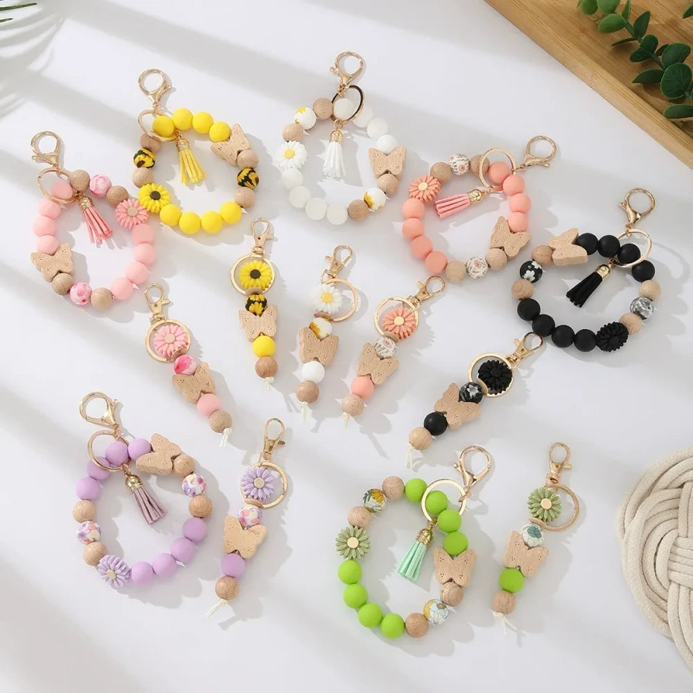 Assorted butterfly and flower silicone and wood bead keychain bracelets in pink, yellow, white, peach, black, purple, and green colorways with gold lobster clasp hardware and tassel accents for DIY jewelry making.