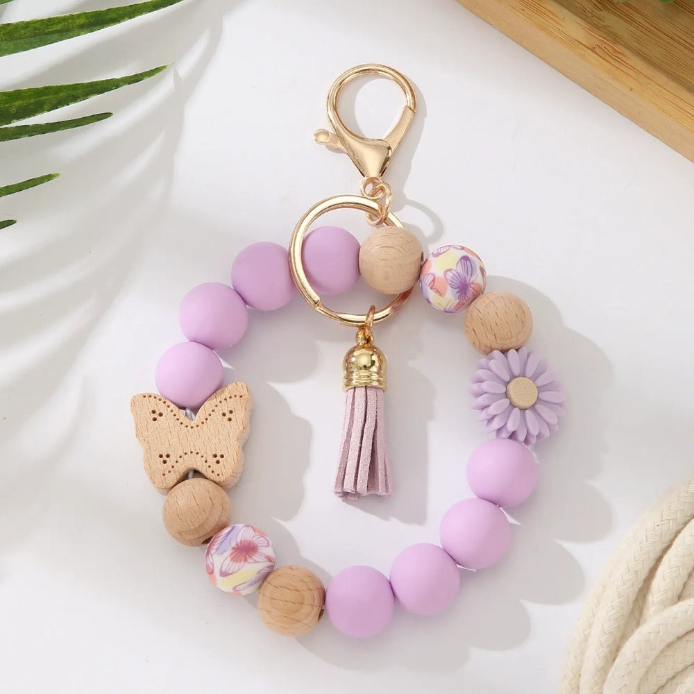 Assorted butterfly and flower silicone and wood bead keychain bracelets in pink, yellow, white, peach, black, purple, and green colorways with gold lobster clasp hardware and tassel accents for DIY jewelry making. - Purple