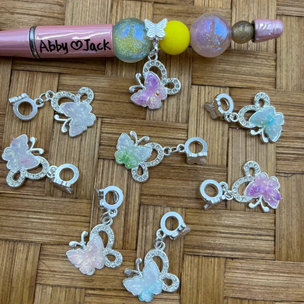 Pastel glitter butterfly rhinestone bail beads in pink, purple, green, and white with silver metal frames, displayed on a wicker surface next to a beaded pink pen labeled Abby and Jack.