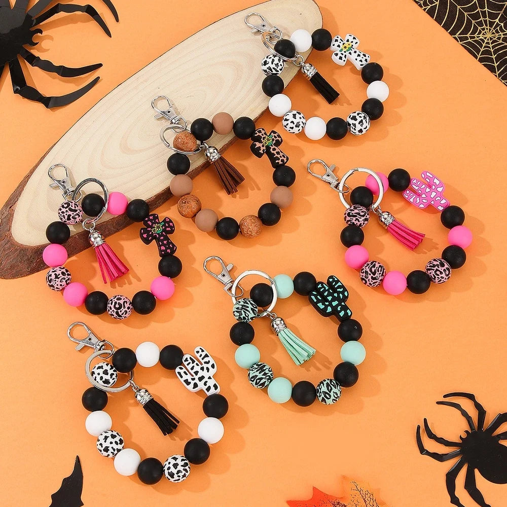 Assorted cactus charm silicone bead keychain bracelets in black, pink, mint, and white with leopard and cow print patterns, displayed on an orange Halloween-themed background.