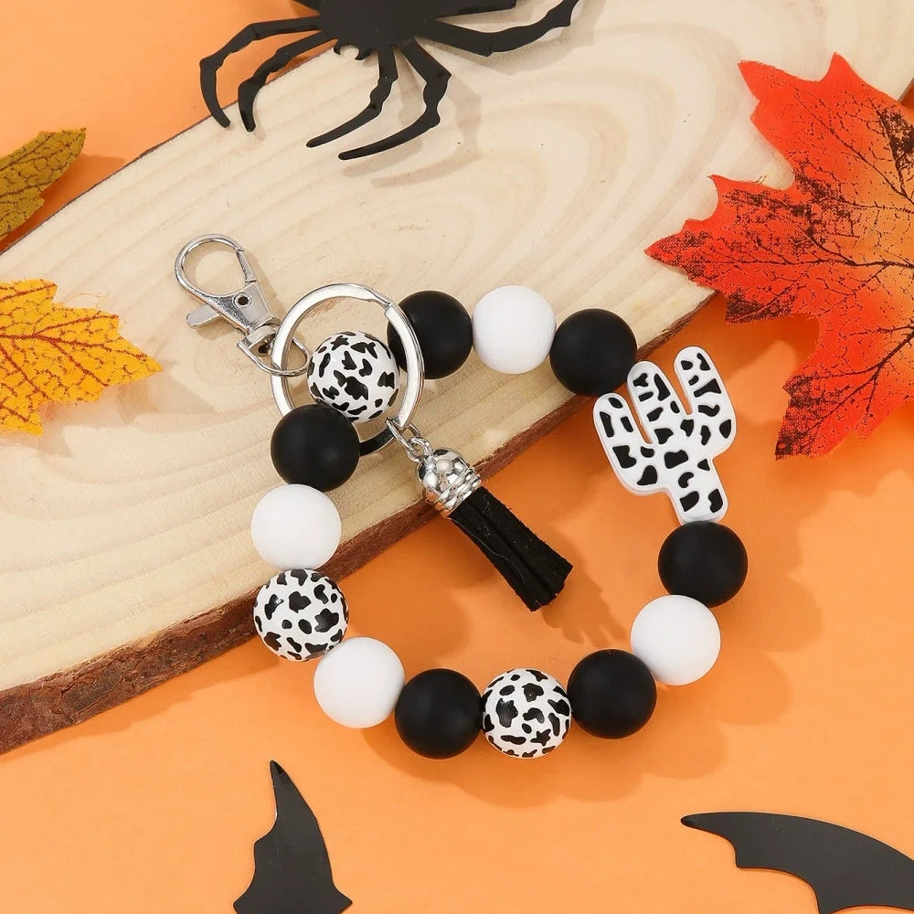 Assorted cactus charm silicone bead keychain bracelets in black, pink, mint, and white with leopard and cow print patterns, displayed on an orange Halloween-themed background. - Black