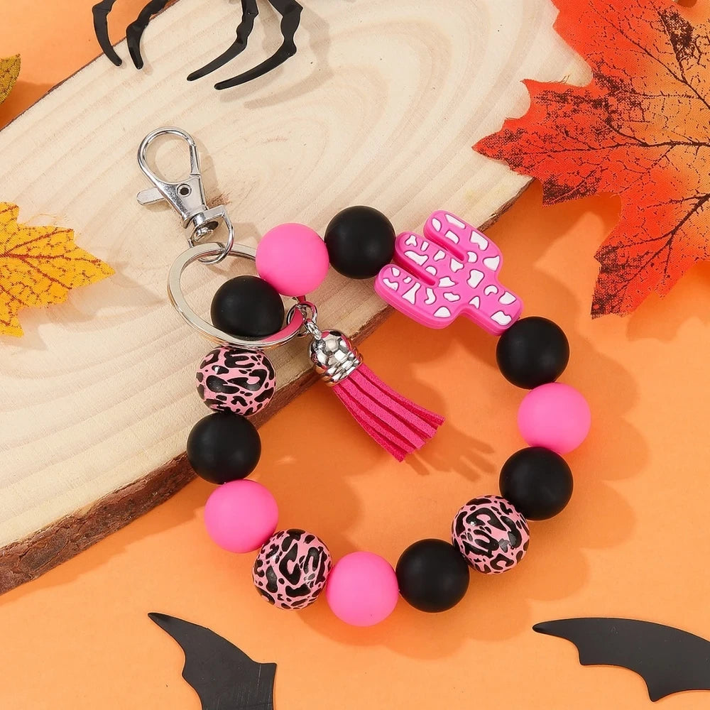 Assorted cactus charm silicone bead keychain bracelets in black, pink, mint, and white with leopard and cow print patterns, displayed on an orange Halloween-themed background. - Pink