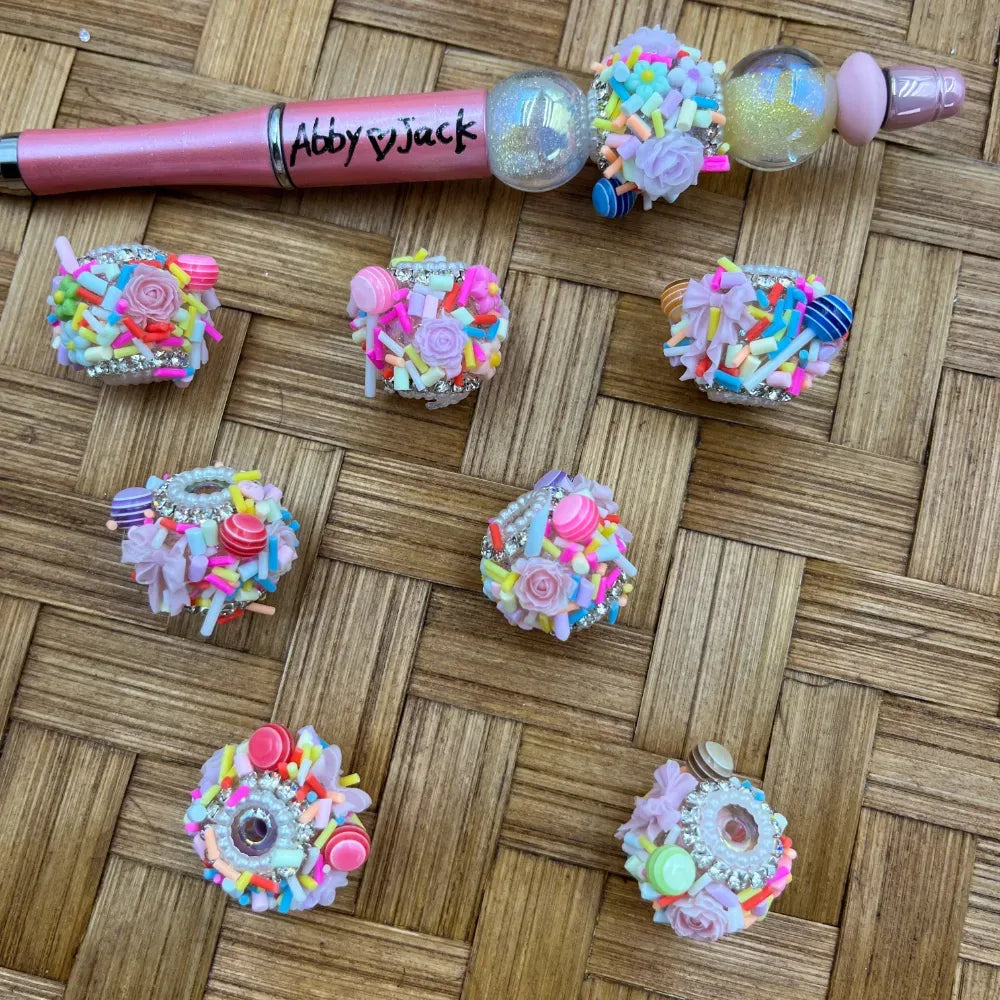 Colorful candy sprinkle and rose rhinestone fancy beads decorated with polymer clay sprinkles, lollipop and floral accents, displayed next to a decorated beadable pen on a wicker background.