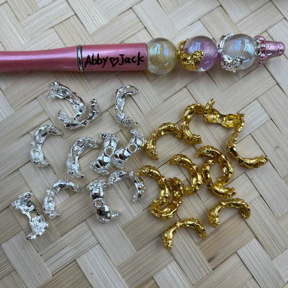 Gold and silver cat-shaped metal bead caps displayed next to a pink beadable pen with decorative beads on a wicker surface.