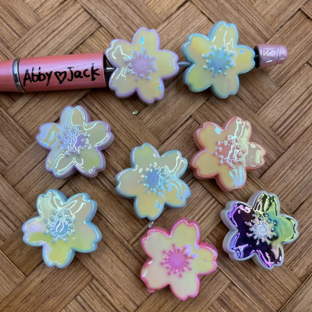 Iridescent cherry blossom flower acrylic beads in pastel yellow, pink, lavender, and dark purple holographic finishes displayed on a woven surface next to a beadable pen labeled Abby loves Jack.