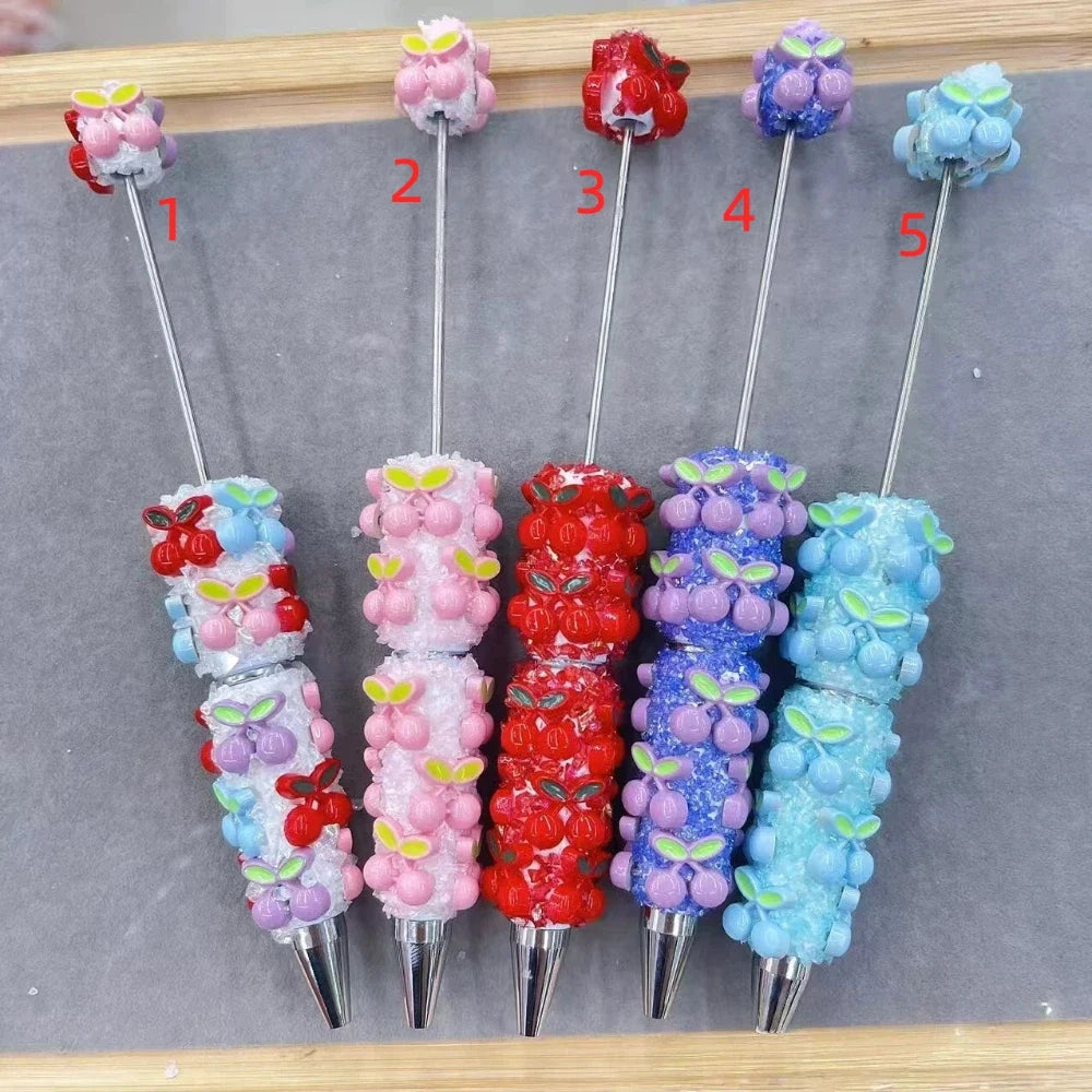 Five decorative beaded clay pens numbered 1-5 featuring cherry cluster charms and sparkle resin crystals in mixed pastel blue, pink, red, purple, and light blue colorways, displayed on a gray velvet surface.