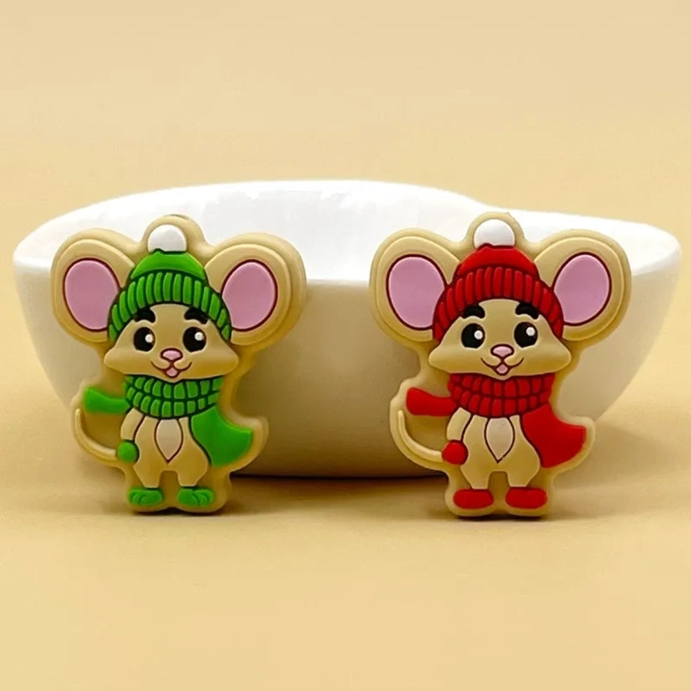 Two cartoon mouse silicone focal beads wearing Christmas winter hats and scarves, one in green and one in red, displayed in front of a white bowl on a beige background.
