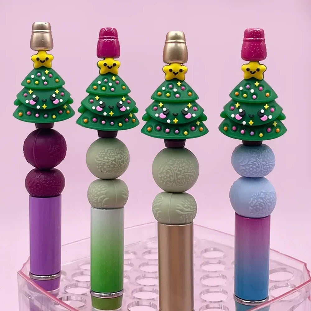 Four Christmas tree 3D silicone focal beads with smiling star toppers, colorful ornament details, displayed on beadable pens in purple, green, gold, and blue color schemes against a pink background.
