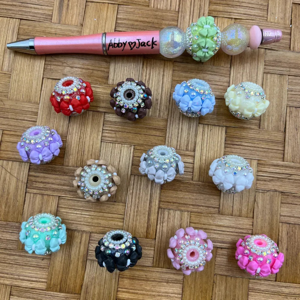 Assorted chunky resin fancy beads decorated with mini bows and rhinestone crystal accents in multiple colors including red, black, pink, lavender, blue, mint, yellow, and white, displayed next to a decorated beadable pen on a wicker surface.